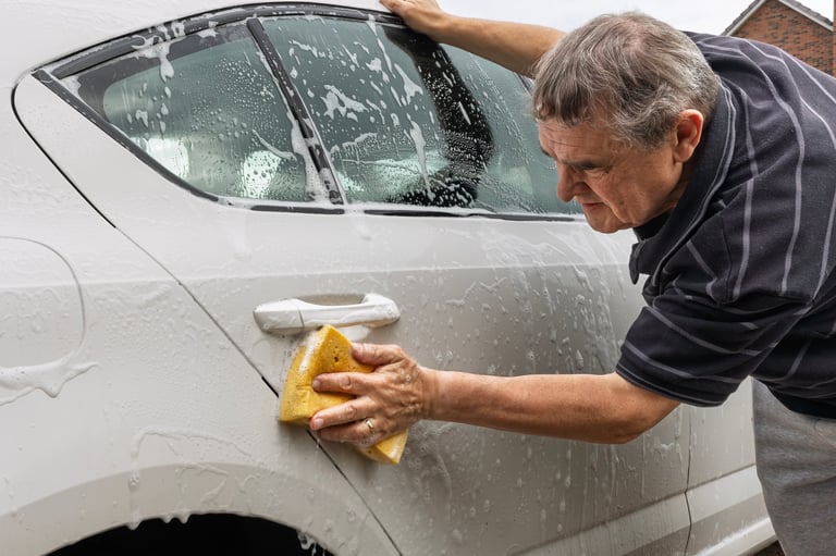 Mature man cleaning the side door of a white car using a yellow sponge and soapy water outside his suburban home